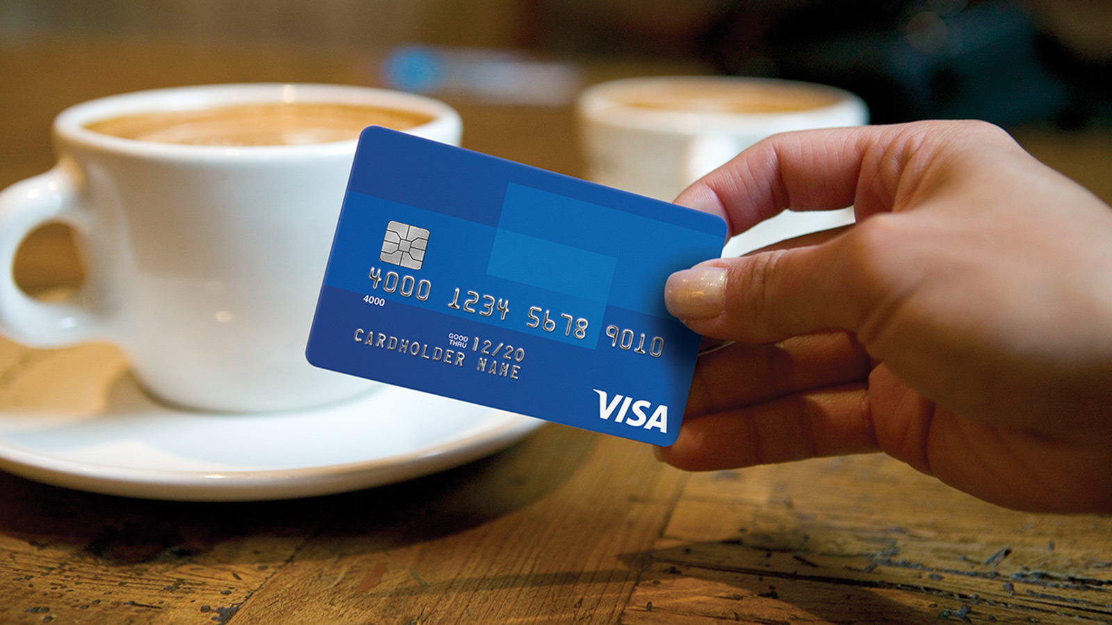 Mão segurando um cartão Visa azul, com uma xícara de café ao fundo